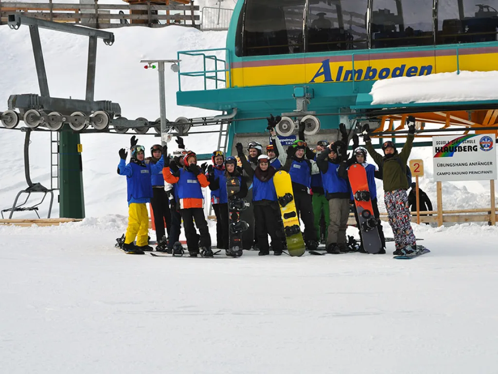 Gruppe posiert mit Snowboards unter dem Schild der Bergstation Almboden.